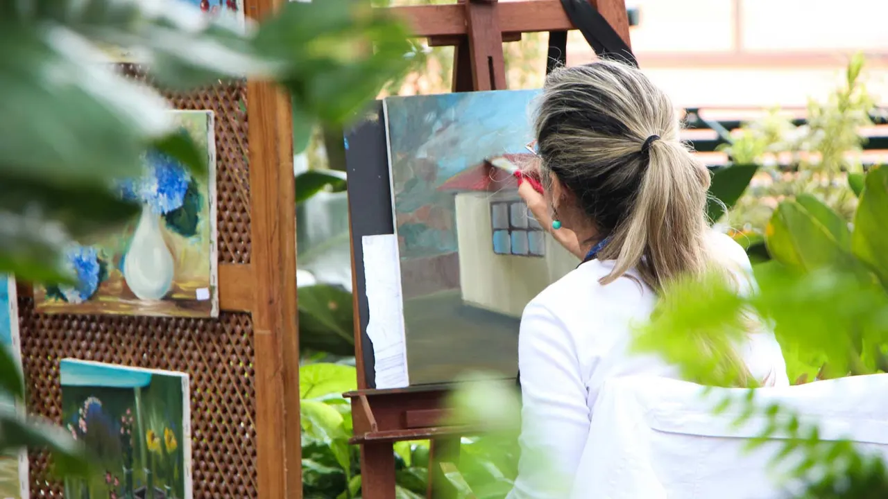 Woman painting at an easel outdoors in a garden surrounded by greenery and flowers.