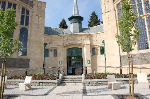 Historic stone building with central entrance, spire, ivy-covered walls, young trees and flower beds in courtyard.