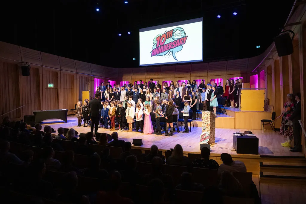 Large group of performers and guests on theatre stage during evening celebration event with audience in foreground.