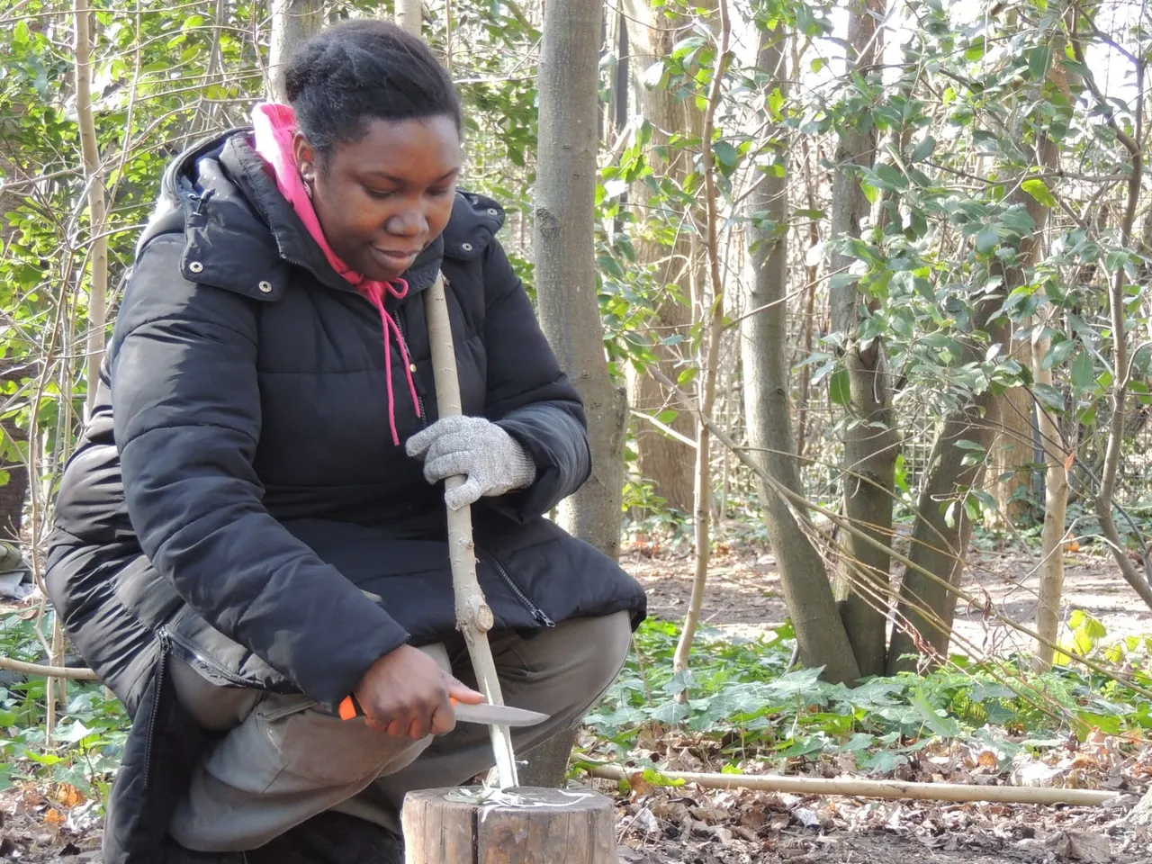 A woman in a black padded jacket crouches whilst using a spade to dig in woodland soil surrounded by bare trees and undergrowth.
