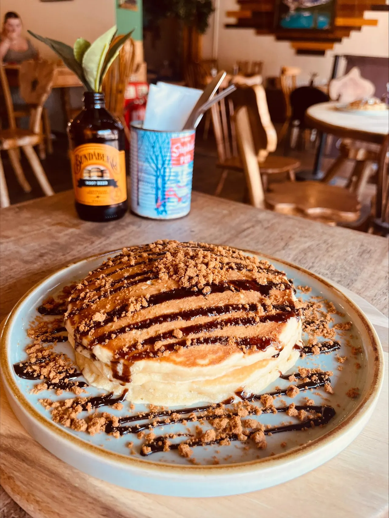 A dessert plate with a cream and chocolate drizzle topped with crumble sits on a wooden table in a dining room with beer bottle and utensil holder nearby.