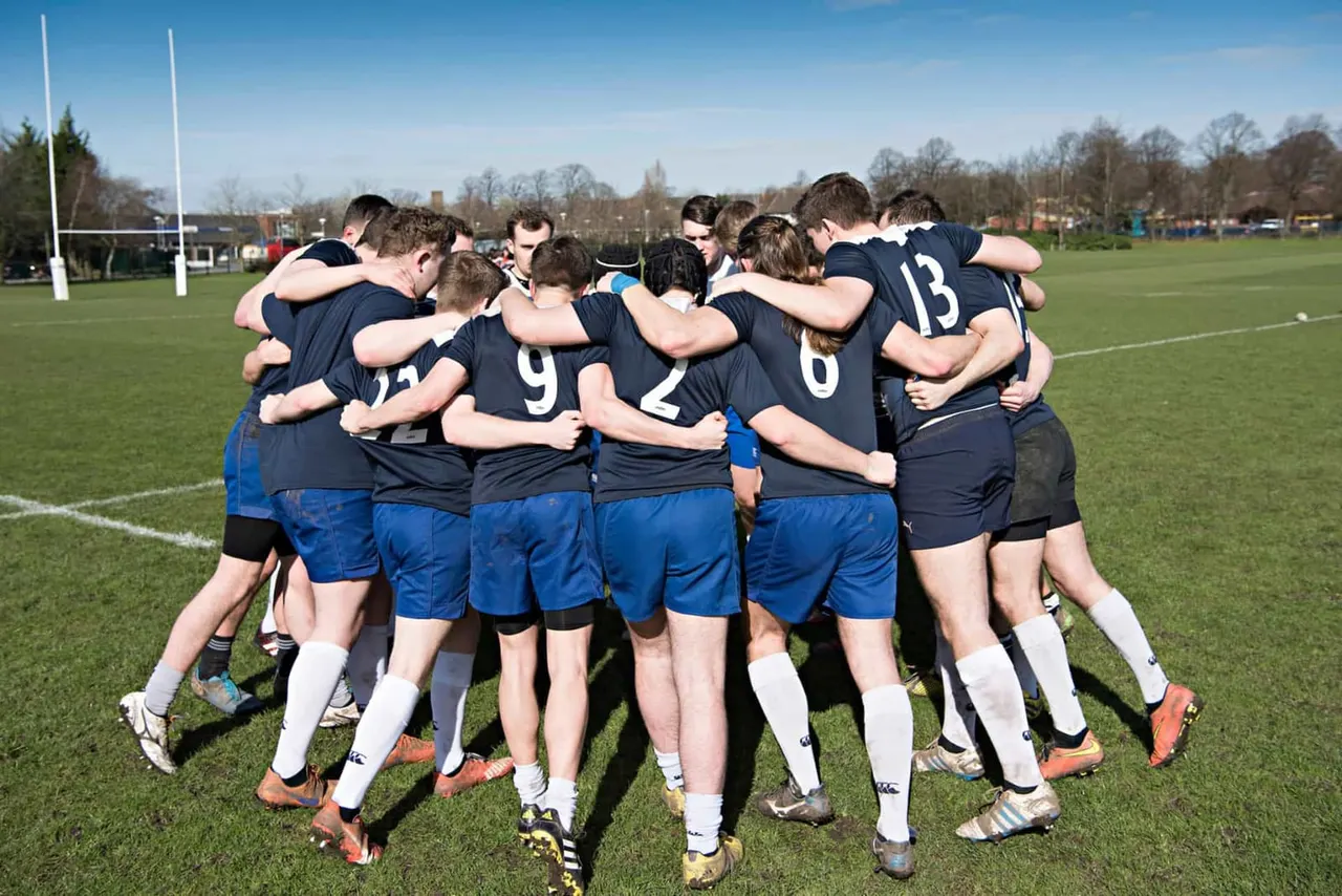 Rugby team in navy and blue kit huddled together on pitch with goal posts and trees in background.