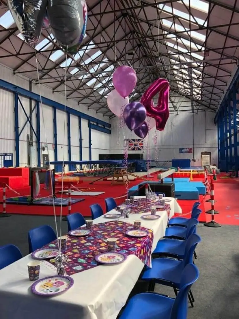 Indoor gymnastics facility decorated for a children's party, with decorated tables, purple balloons, and padded play equipment visible in the background.