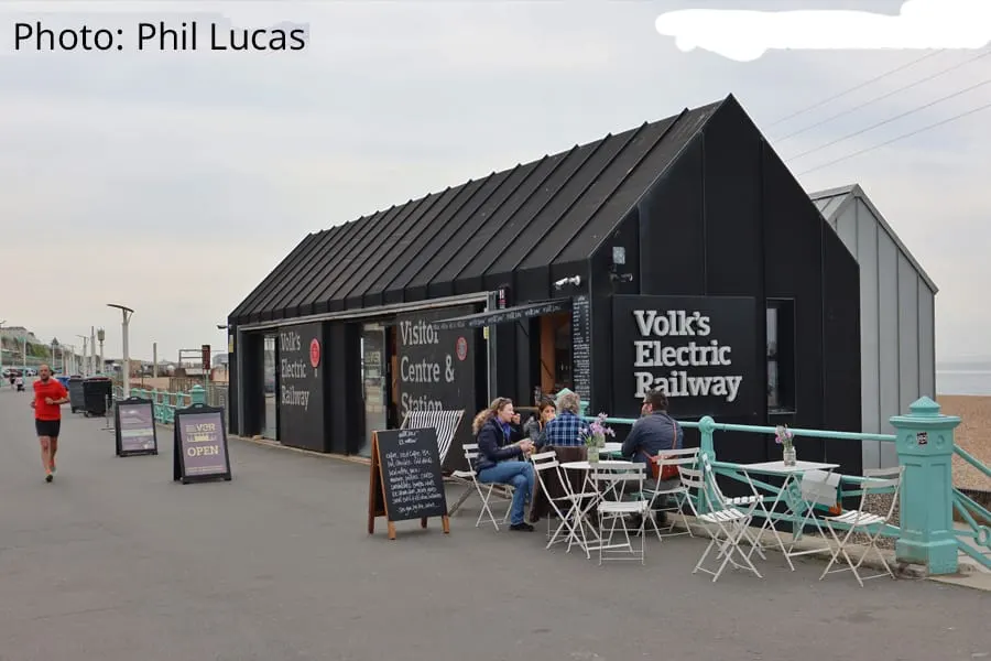 Visitors seated at outdoor tables outside a black building with "Volk's Electric Railway" signage on a beachside promenade.