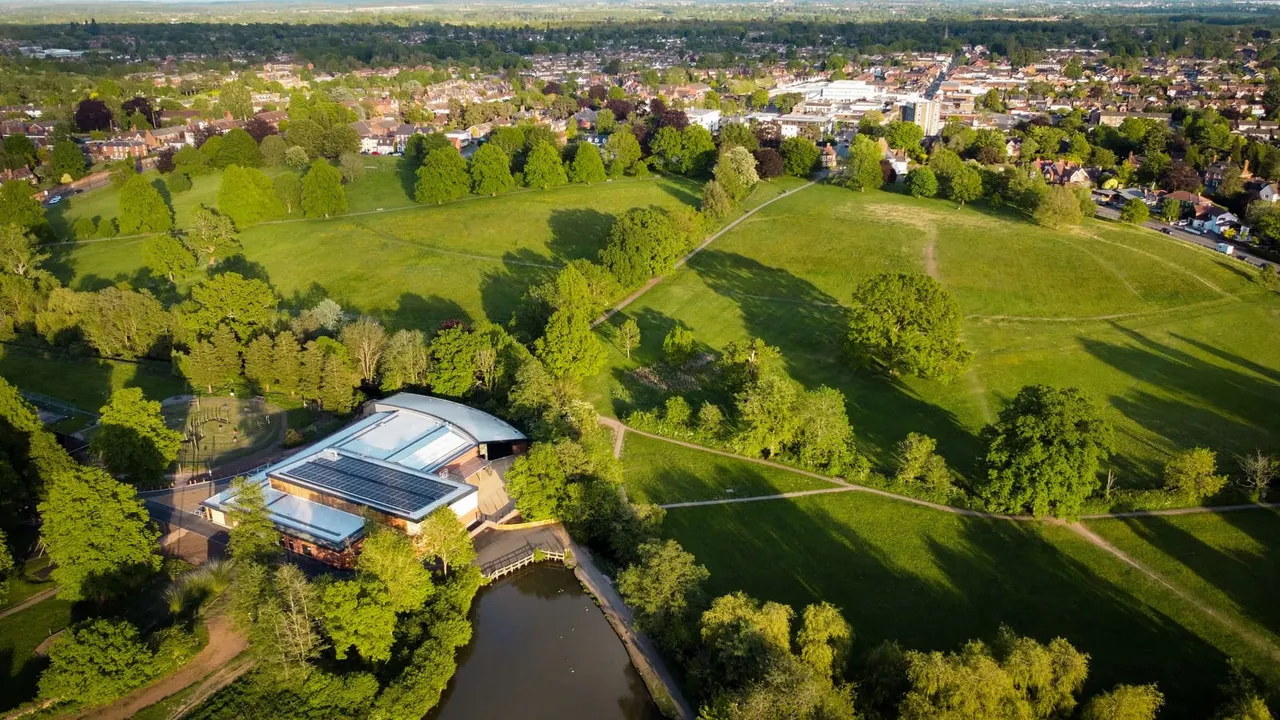 Aerial view of a modern leisure building with solar panels beside a river, surrounded by parkland, trees and suburban housing.