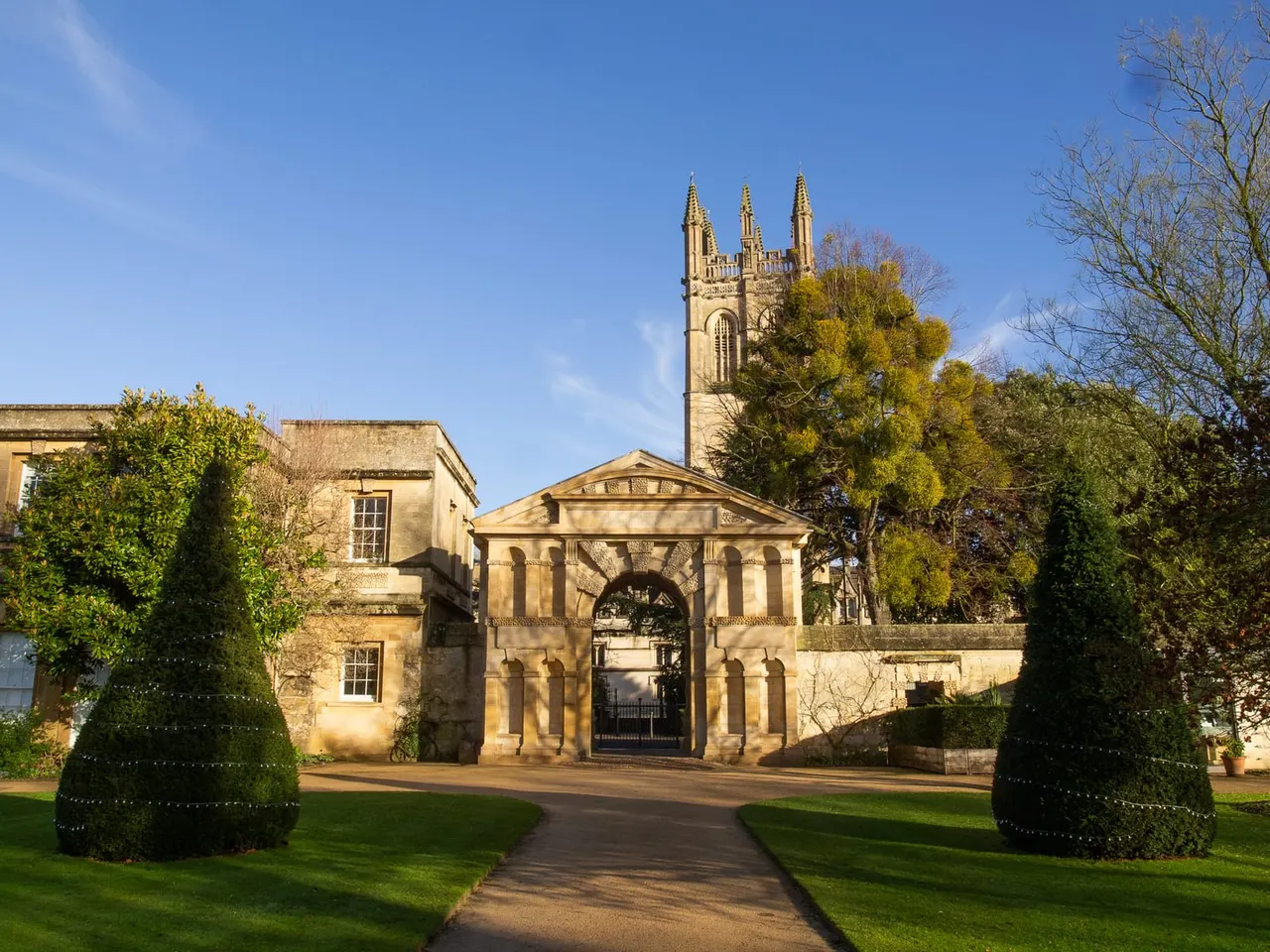 Historic stone gatehouse and church with spire viewed across manicured lawns and topiary in autumn sunlight.
