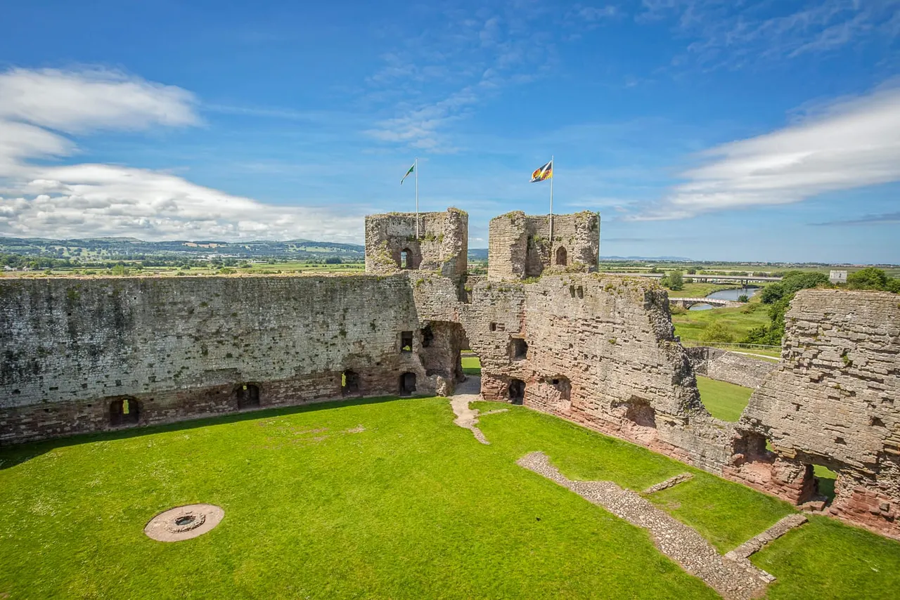 Interior courtyard of a medieval castle with stone towers, flags, and green lawn, overlooking countryside.