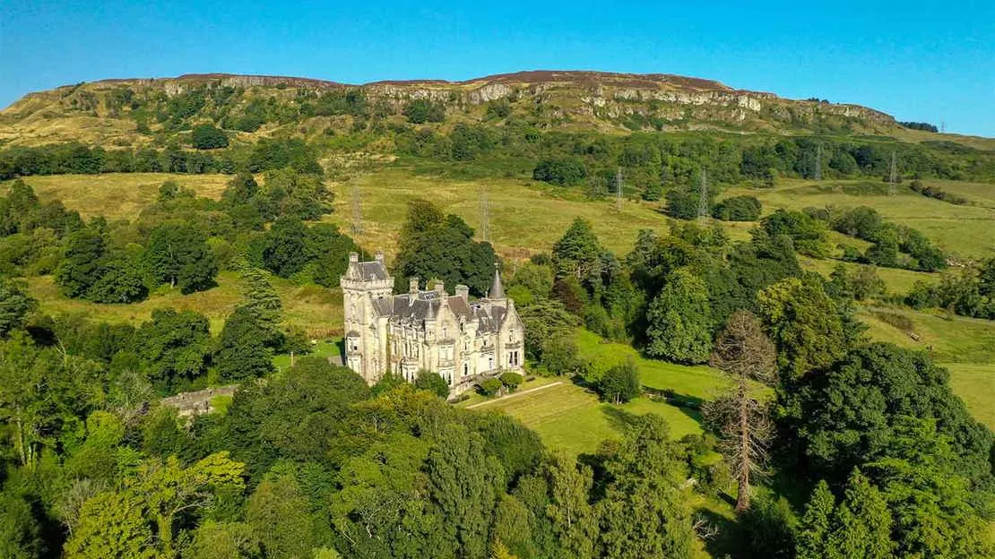 Stone castle nestled in lush green woodland with hills in the background under clear blue sky.