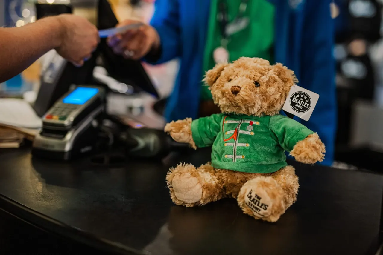 A teddy bear wearing a green Beatles jacket sits on a counter whilst staff process a payment behind it.