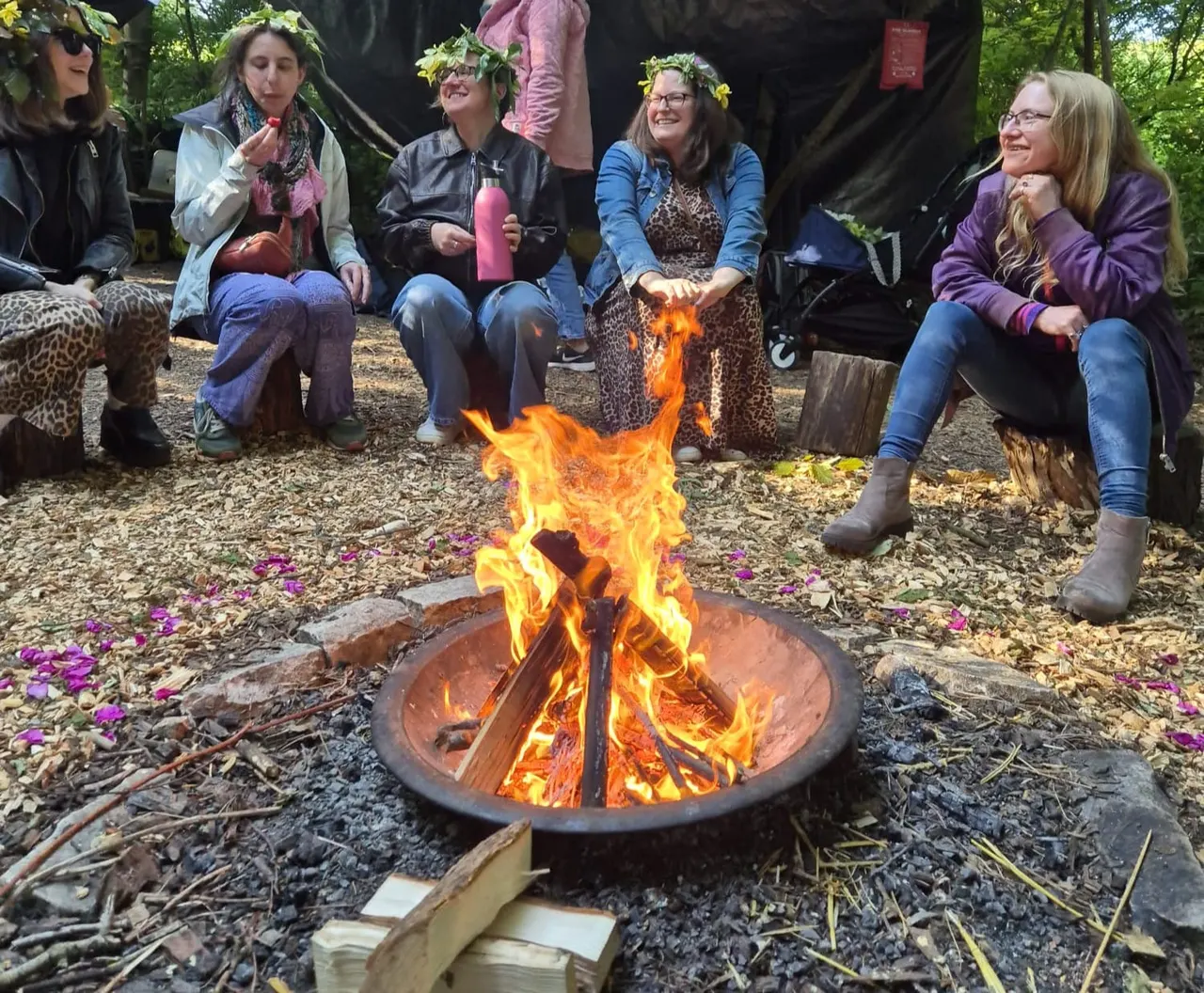 Group of smiling visitors seated around a blazing campfire in a woodland clearing, wearing flower crowns.