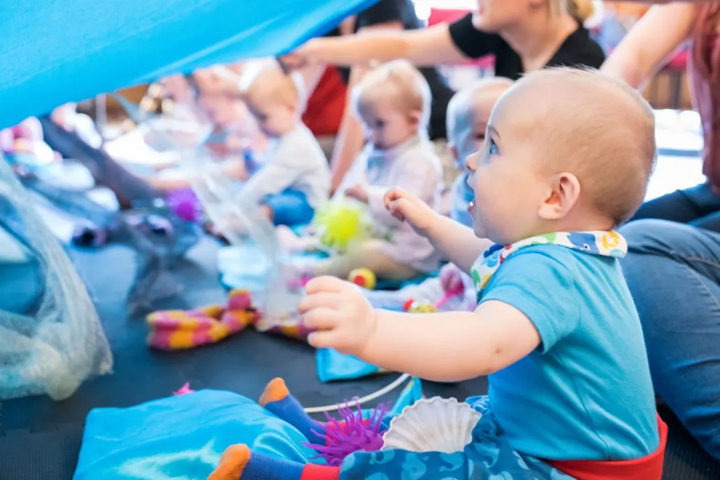 Toddler in blue shirt playing with colourful sensory toys in a ball pool with other young children and carers.