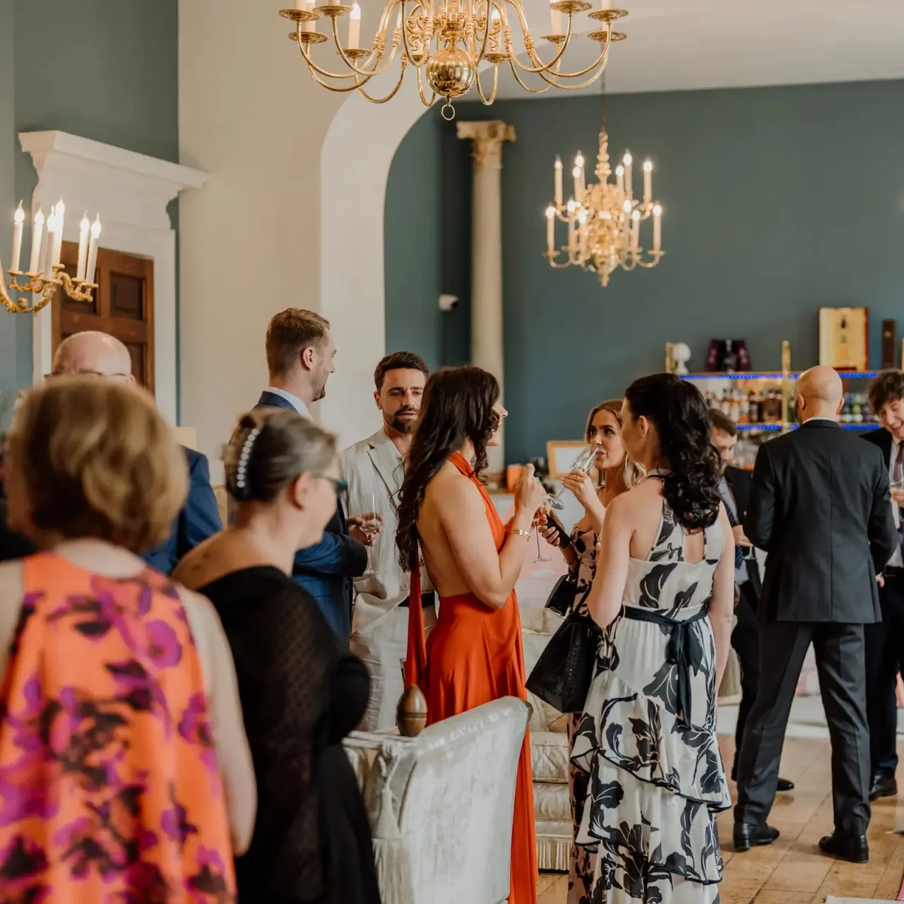 Guests in formal attire mingle and hold drinks in an elegant room with green walls, gold chandeliers, and classical architectural details.