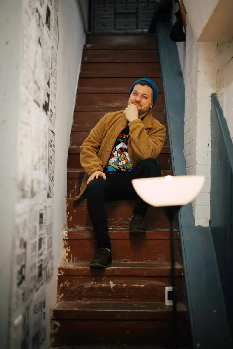 Man in blue beanie and mustard cardigan sits pensively on wooden stairs in a narrow stairwell.