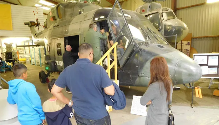 Visitors boarding a military helicopter on display in an indoor hangar museum.