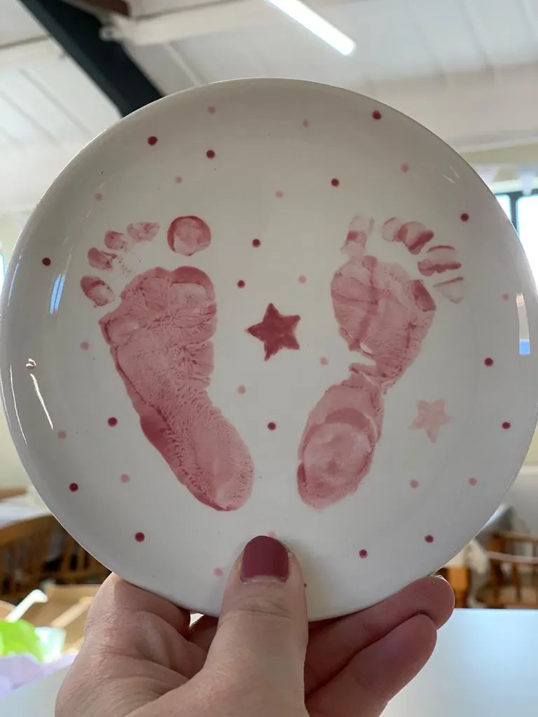 A hand holds a white ceramic plate decorated with pink baby footprints and stars.