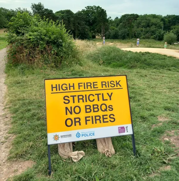 A yellow "High Fire Risk" warning sign in a grassy field with trees and a path in the background.