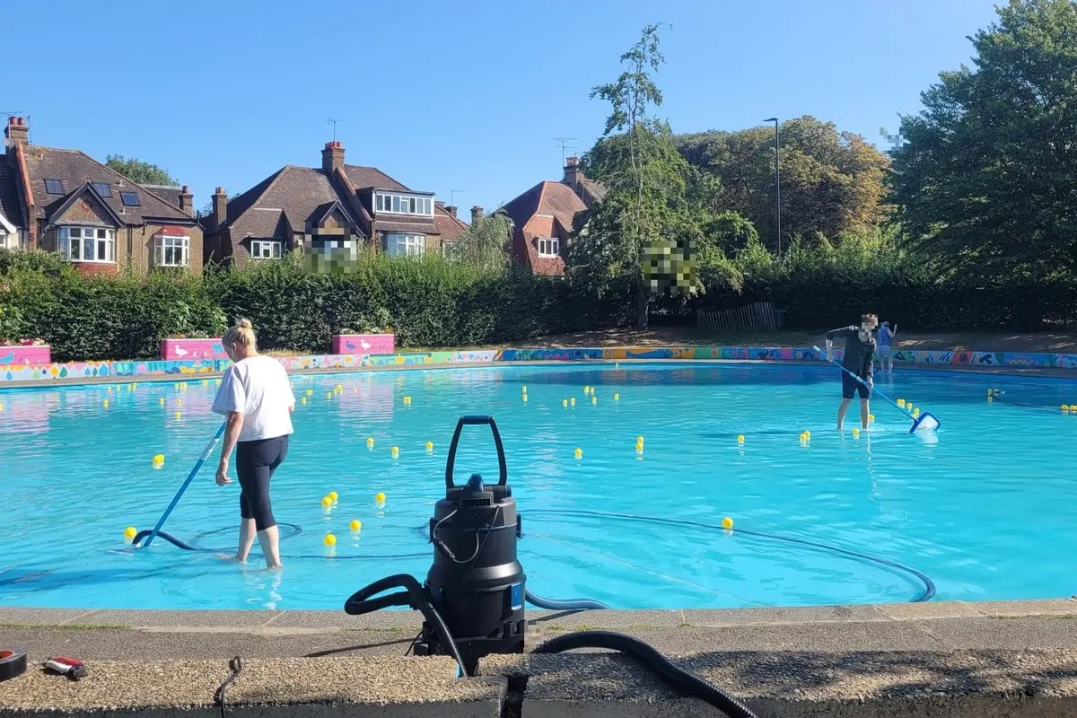 Staff member and children in a bright turquoise outdoor swimming pool with yellow lane markers on a sunny day.