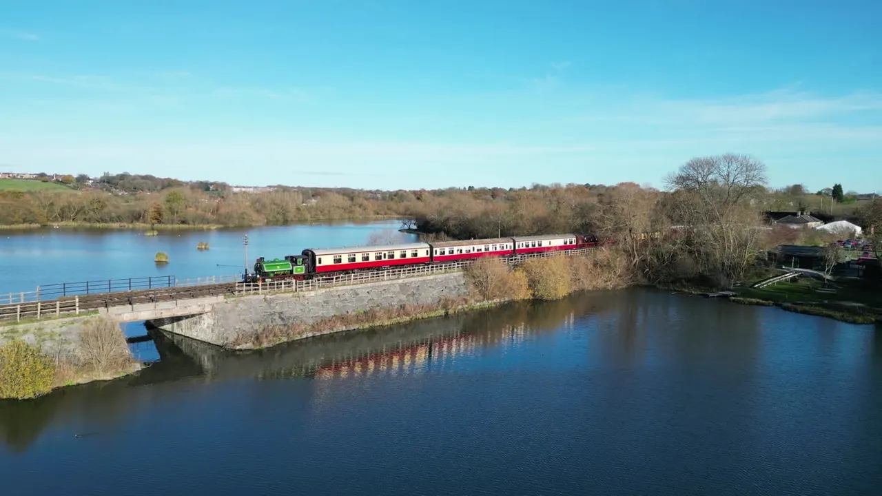 Steam train with red and white carriages travelling along track beside canal and wooded countryside.