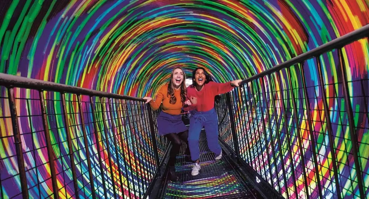 Two visitors walk through a vibrant rainbow light tunnel with concentric circular neon colours.