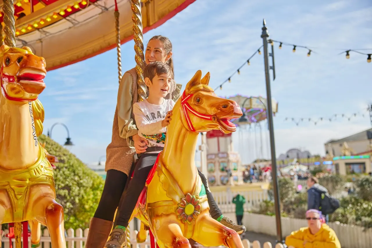 An adult and child ride a golden carousel horse at a fairground, with rides and attractions visible in the background.