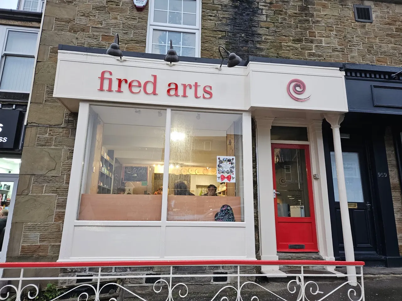 Storefront of a pottery and ceramics studio with white fascia, large windows displaying work, and a red entrance door in a stone-built street.