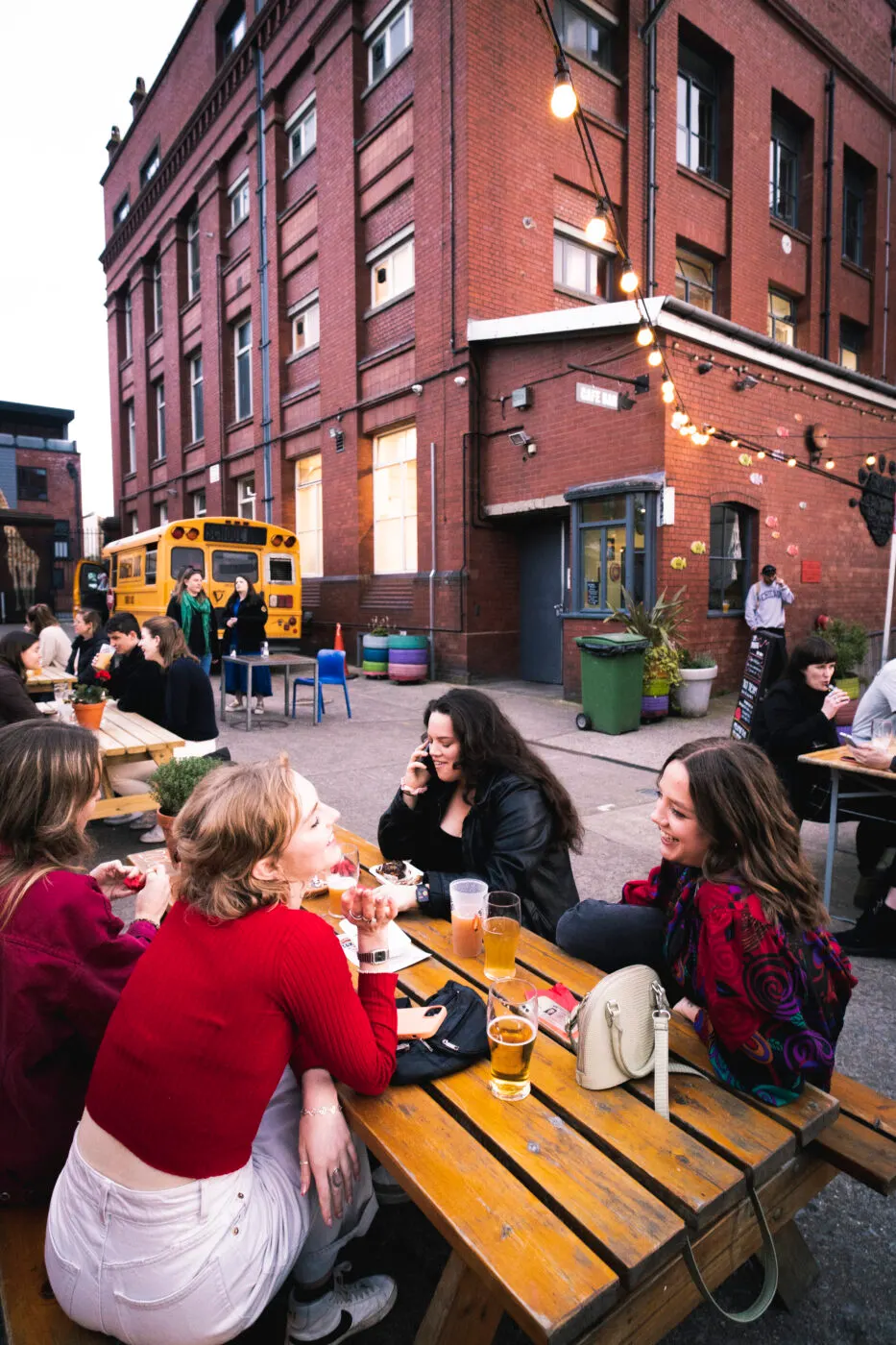 People dining at wooden tables in a brick courtyard with a yellow vehicle and string lights overhead.