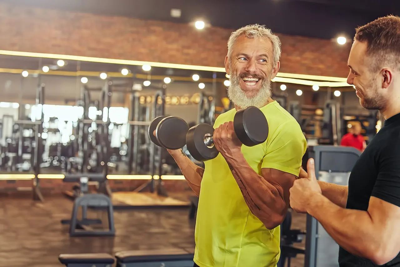 A smiling man in a lime-green t-shirt lifts dumbbells whilst chatting with a trainer in a modern gym.