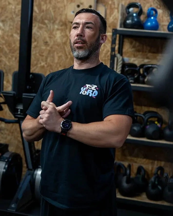Man in black t-shirt speaking in a gym surrounded by kettlebells and exercise equipment on shelves.