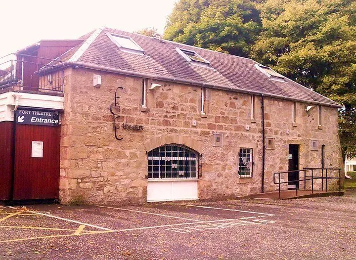 Red sandstone theatre building with purple roof and entrance door, surrounded by trees.