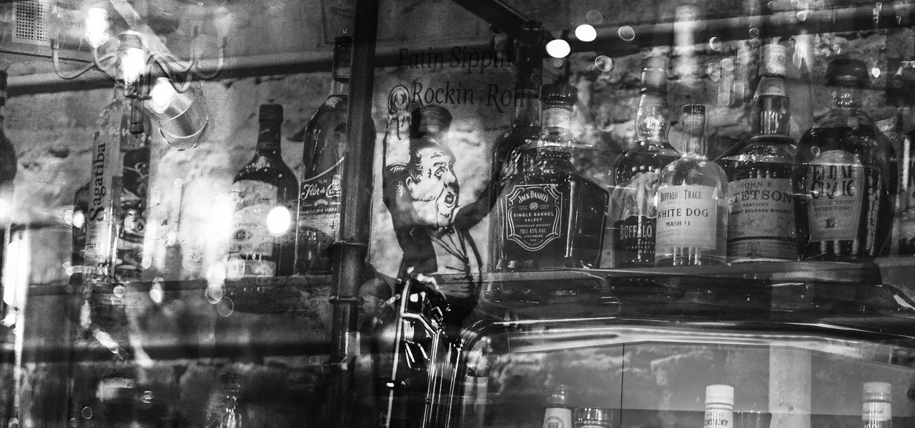 Black and white photograph of a bar window display with bottles of spirits and vintage neon signage reflected in the glass.