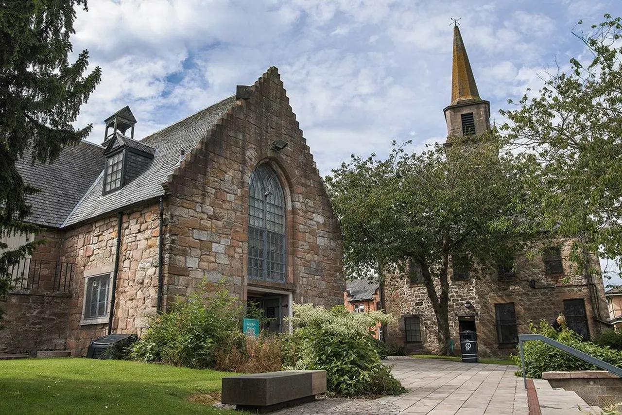 Historic stone church building with pitched roof and golden spire, surrounded by green lawn and mature trees on a sunny day.