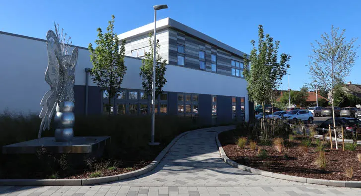 Modern leisure centre building with curved entrance pathway, young trees, and sculpture feature in foreground.