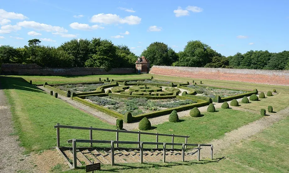 Formal parterre garden with low hedging and topiary cones, brick walls visible beyond, under blue sky.
