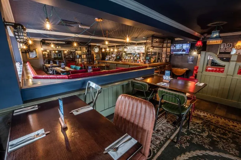 Interior of a pub with wooden flooring, dining tables, chairs, and a bar with red neon trim and shelved bottles.