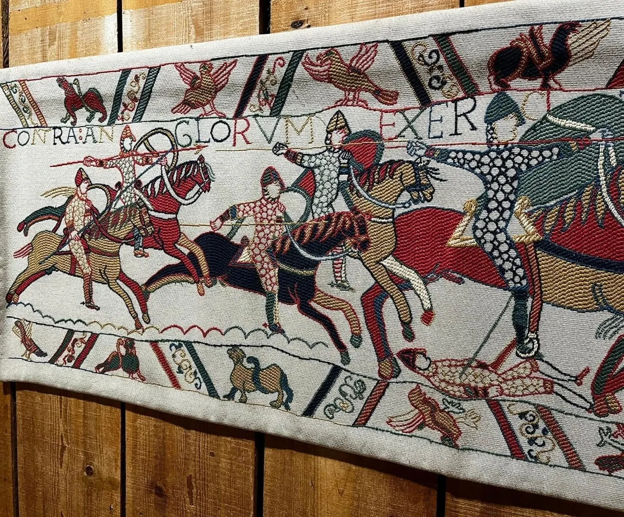 Medieval embroidered tapestry depicting Norman warriors on horseback and foot soldiers in battle scenes against wooden boards.
