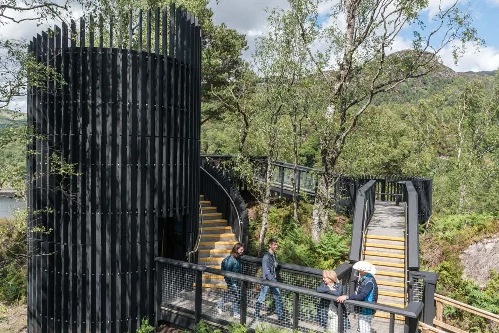 Visitors walk through a modern black wooden structure with yellow-stepped walkways surrounded by trees and water in a highland landscape.