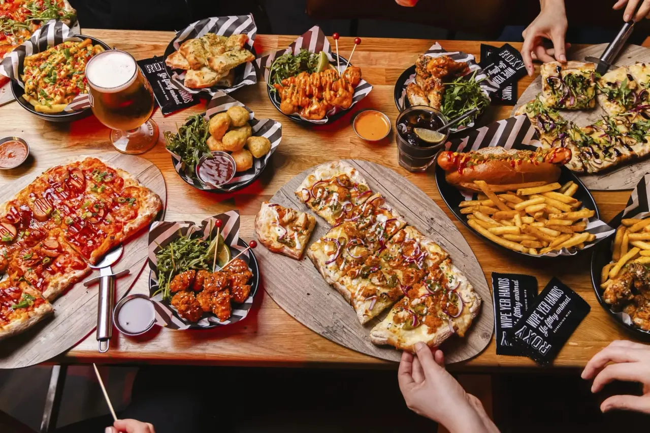Group sharing pizza, hot dogs, fried chicken, chips and other pub food at a wooden table with beer.
