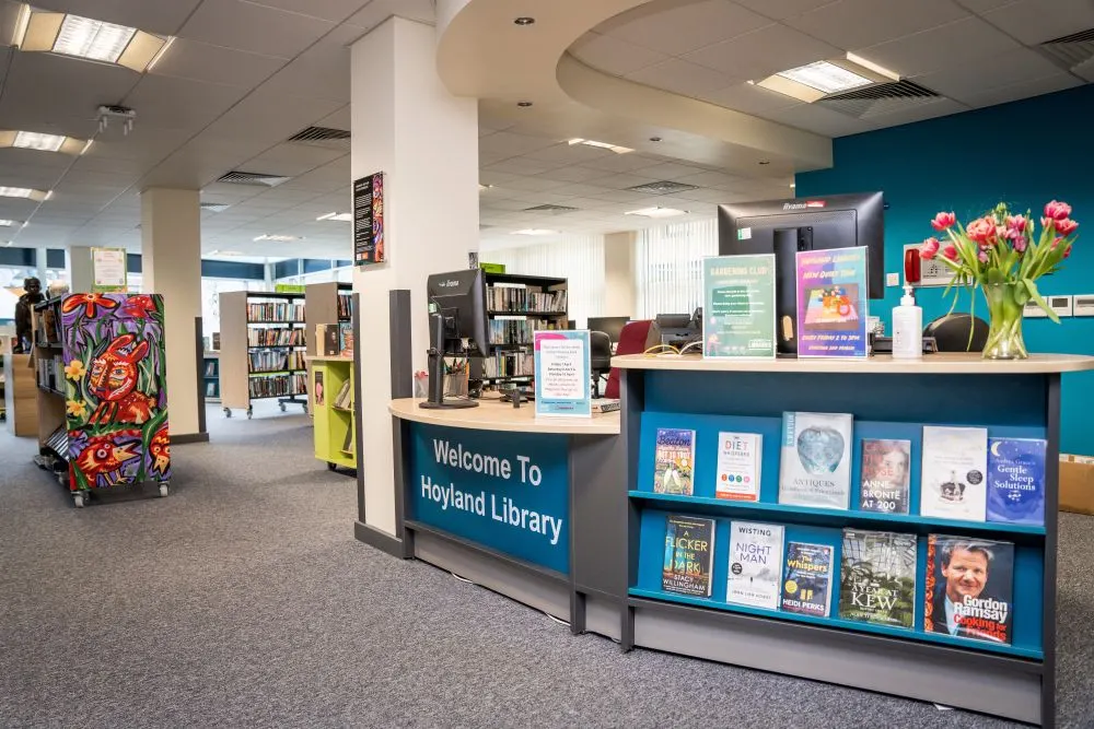 Interior of a public library with reception desk, bookshelves, and colourful artwork on display columns.