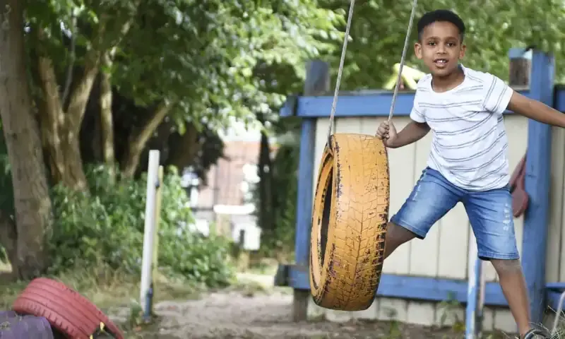 Child playing on a tyre swing in an outdoor play area with trees and blue structures.