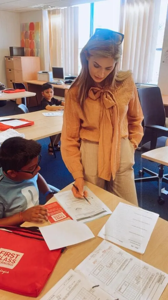 An instructor in a yellow shirt works with a young child at a desk in a bright classroom, with other children studying at tables in the background.