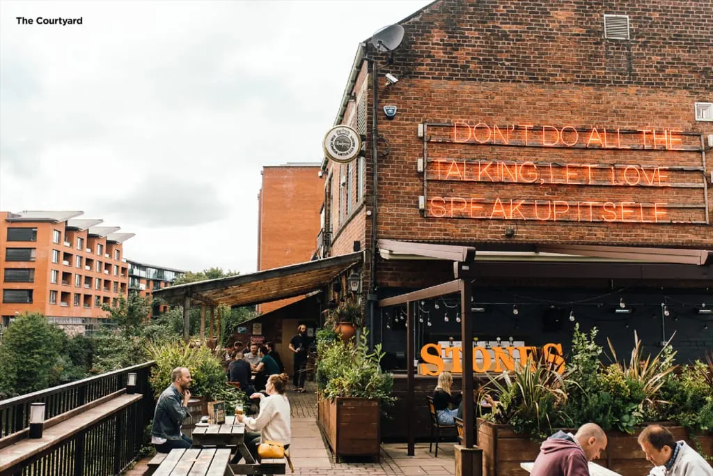 People dining and socialising on a rooftop terrace with wooden decking, potted plants, and a brick building with neon signage.