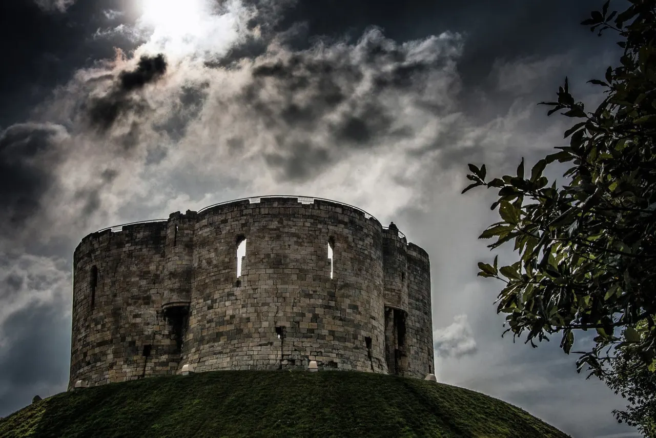 Clifford's Tower, a circular stone keep, sits on a grass mound beneath dramatic cloudy skies with foliage visible on the right.