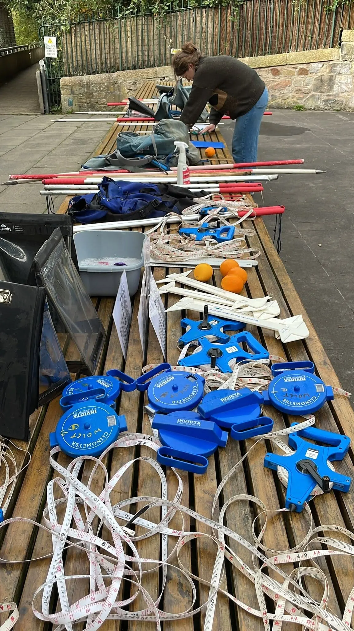 Two people organizing outdoor activity equipment on wooden tables, including skis, climbing gear, harnesses, and blue water sports kit.