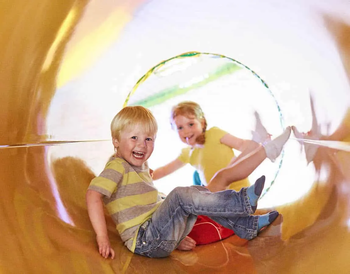 Two young children playing inside a soft play tunnel structure, smiling at the camera.