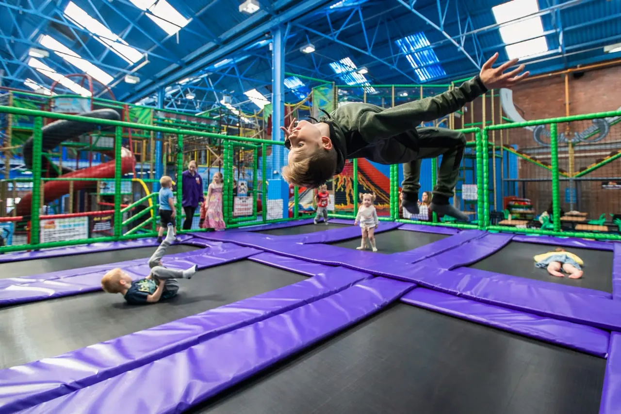 Child doing a backflip on a purple trampoline whilst other children play in an indoor trampoline park.