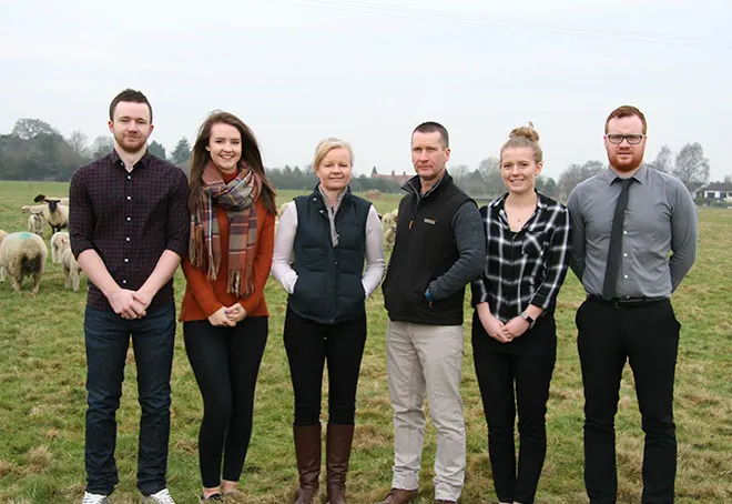 Six people standing in a line in a farm field with sheep grazing in the background.