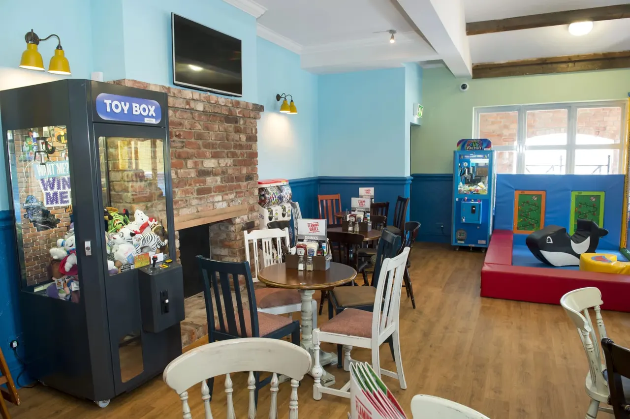 Indoor play area with dining tables, arcade games, a toy box machine, and soft play equipment.