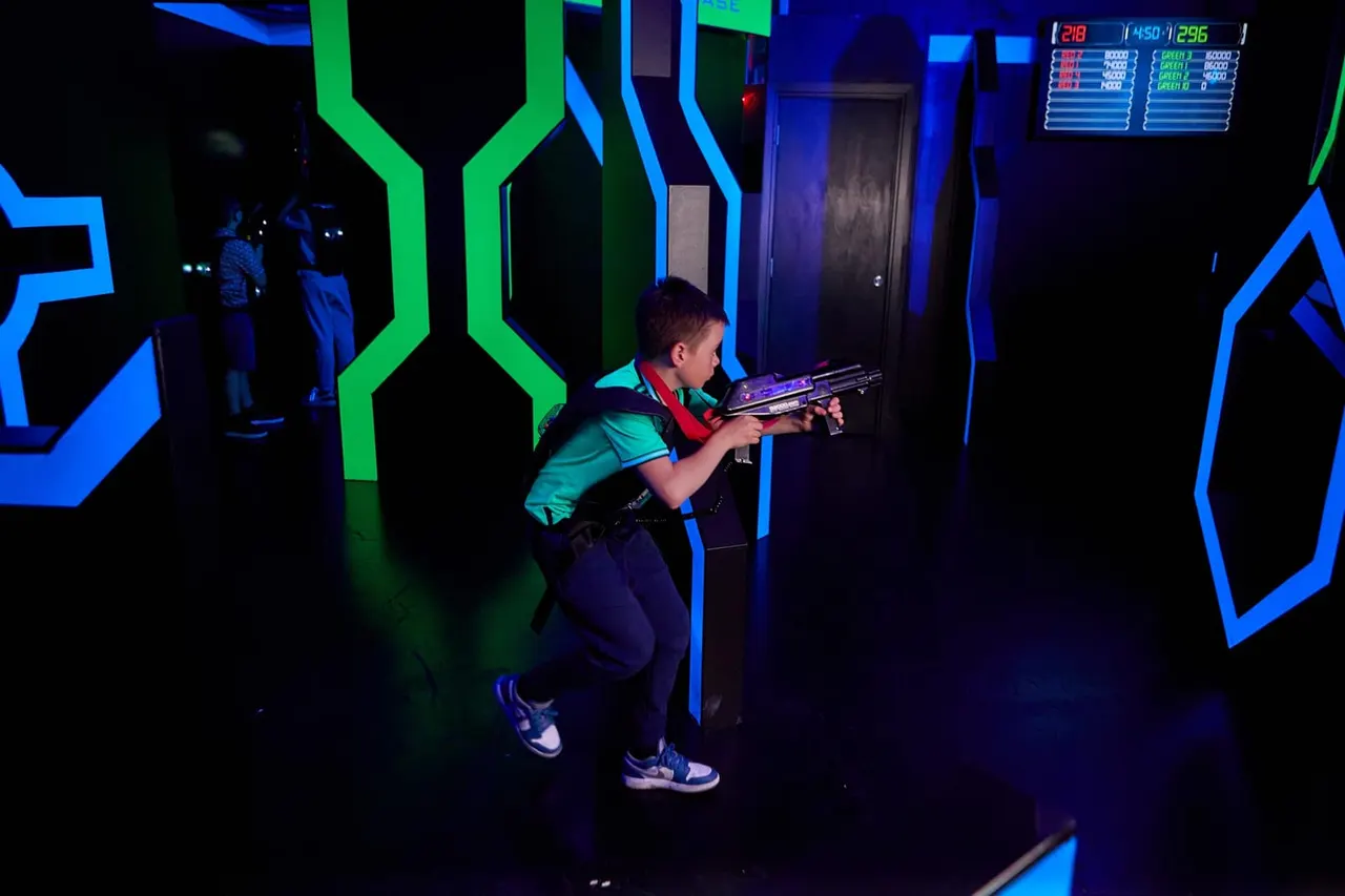 Child playing laser tag in a neon-lit arena with blue and green geometric walls and scoreboards.