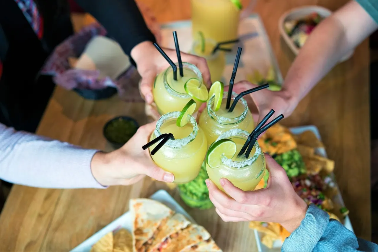 People holding yellow cocktails with lime slices and black straws at a social gathering.