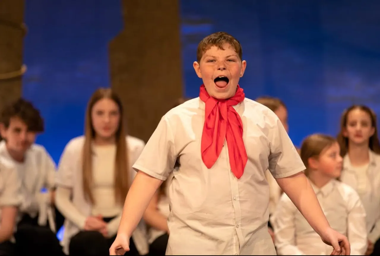 Young boy in Scout uniform with red neckerchief singing on stage with fellow Scouts behind him.